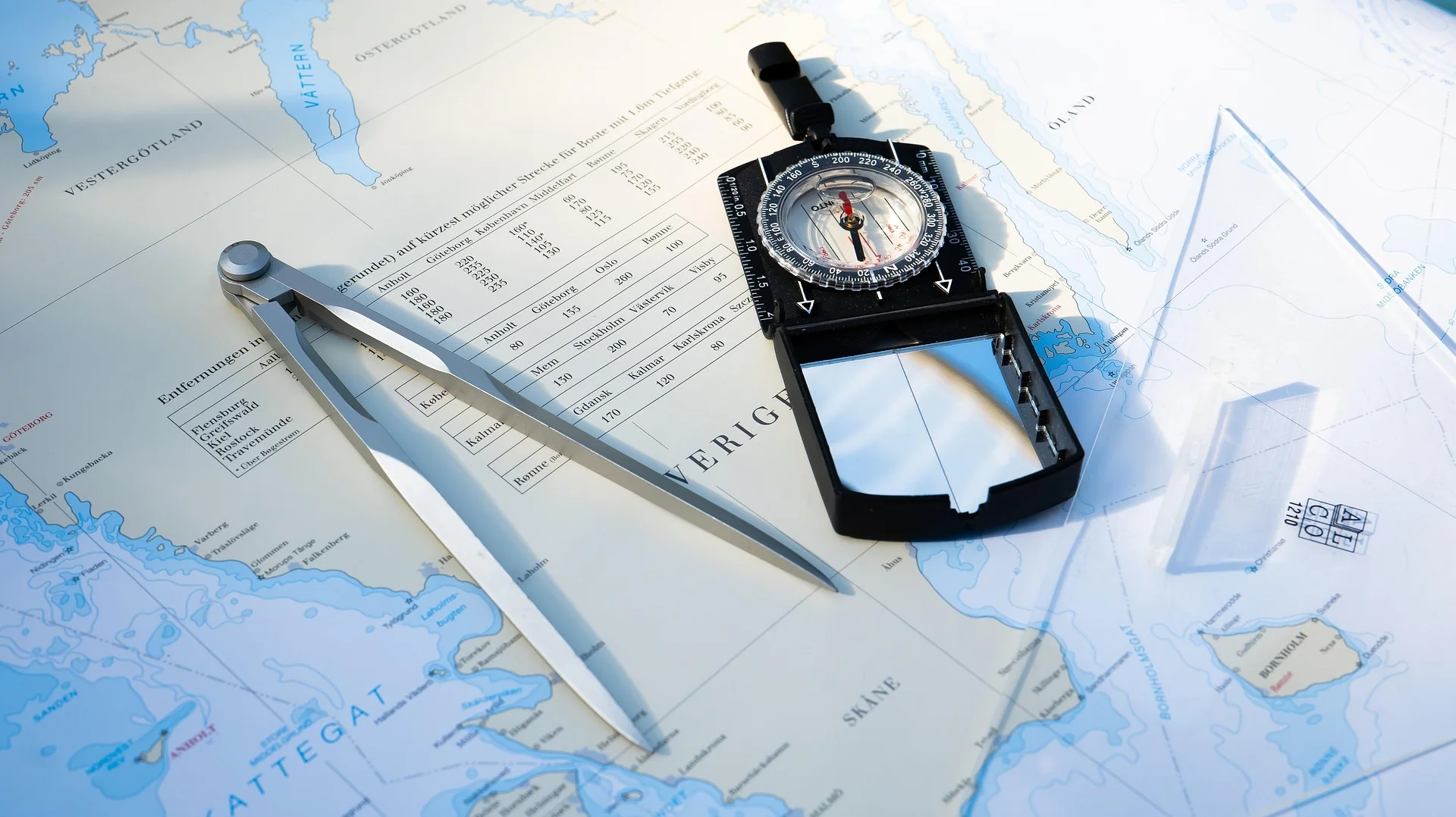 A nautical chart is spread across a table with a navigation compass, divider calipers, and transparent plotting tools arranged on top. The instruments are positioned over coastal and maritime map markings, suggesting route planning or marine navigation. Bright natural light casts soft shadows across the chart.
