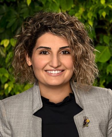 Headshot of Naira Tumanyan, a middle-aged woman with curly light brown hair and blonde highlights, smiling at the camera. She is wearing a gray jacket over a dark navy top, with blurred green foliage in the background.