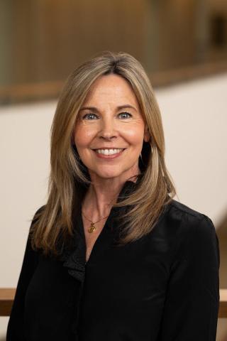 Head shot of Rebecca Wenrich, who is wearing a black blazer and is framed with a blurred brown background. 