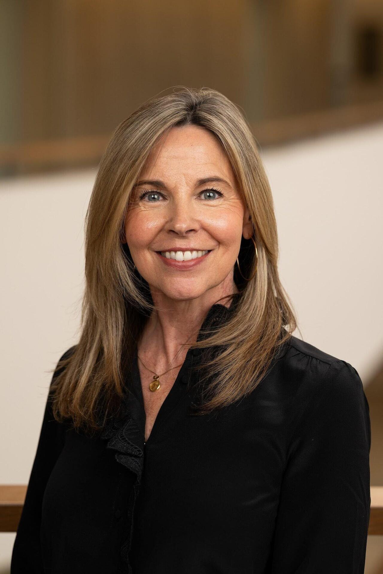 Head shot of Rebecca Wenrich, who is wearing a black blazer and is framed with a blurred brown background. 